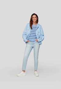 Hellblaues Zip-Up-Jackett über einem gestreiften blauen Pullover, kombiniert mit hellen Jeans und weißen Sneakern. Lässiges Outfit, das für verschiedene Anlässe geeignet ist.