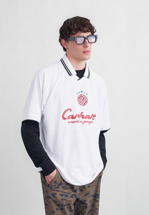 Ung mand med krøllet hår iført oversized sorte briller, hvid polo med rød logo, sorte langærmede trøje og mønstrede bukser, hænderne i lommerne.