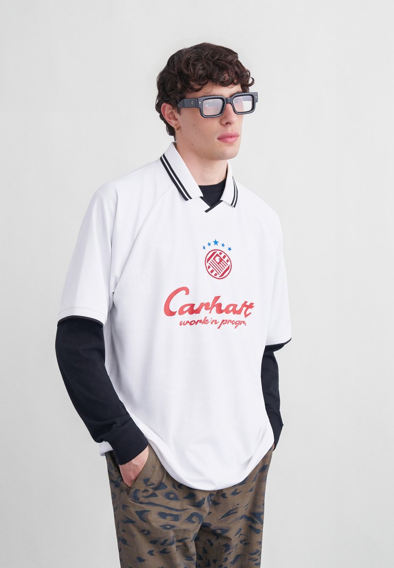 Junger Mann mit lockigem Haar, der übergroße schwarze Brillen, ein weißes Poloshirt mit rotem Logo, schwarze Langarmshirts und gemusterte Hosen trägt, die Hände in den Taschen.