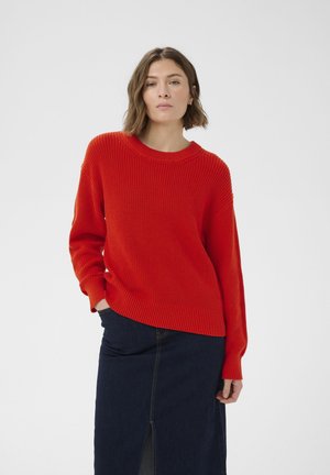 Rövid barna hajú nő piros kötött pulóvert és sötétkék farmer szoknyát visel, amelynek elején hasíték található, és egyszínű fehér háttér előtt pózol.