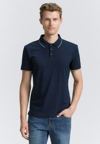 Marineblauwe poloshirt met een kraag en drie knopen, met contrasterende lichte afwerking op de kraag en mouwen, gecombineerd met een blauwe spijkerbroek.