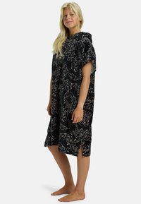 Poncho con capucha en tela oscura con patrones florales en beige. Presenta mangas cortas y aberturas laterales para facilitar el movimiento. Textura suave.