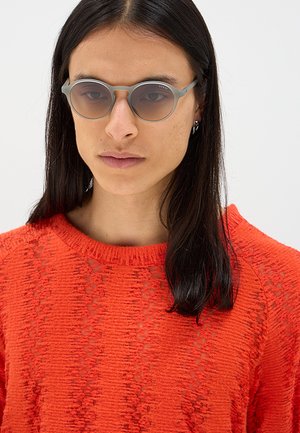 Persona con capelli lunghi e scuri che indossa occhiali da sole rotondi grigi e un maglione a maglia arancione acceso e strutturato su uno sfondo neutro.