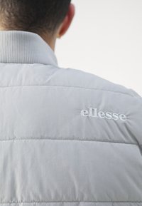 Ljust grått vadderat jacka med ribbat krage, med den broderade logotypen "ellesse" på baksidan. Slät textur, modern design.