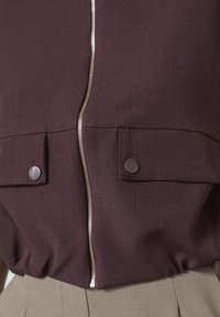 Bordeauxjakke med sølvlynlås, som har to lapperlommer med trykknaplukninger og en gathered kant. Stoffet har en tekstureret overflade.