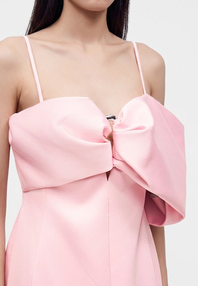 Femme portant une robe rose clair avec de fines bretelles et un grand nœud au niveau de la poitrine maintenu par un anneau argenté.