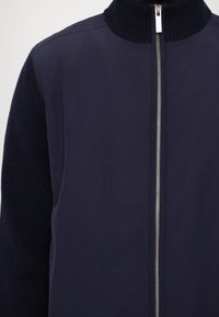 Navy jacket with a ribbed high collar, featuring a full zip front and textured fabric on the sleeves. Smooth, lightweight material.
