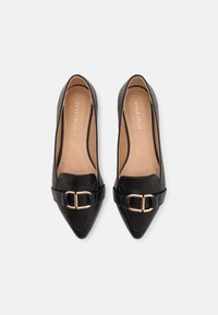 Ballerines noires en cuir avec un bout pointu, ornées d'une boucle dorée et d'une texture lisse. L'intérieur est doublé de cuir tan avec la marque "ANNA FIELD".