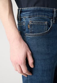 Blugi din denim albastru închis cu cusături portocalii și o etichetă mică "BOSS" pe buzunarul din spate. Prim-plan al mâinii odihnindu-se pe material.