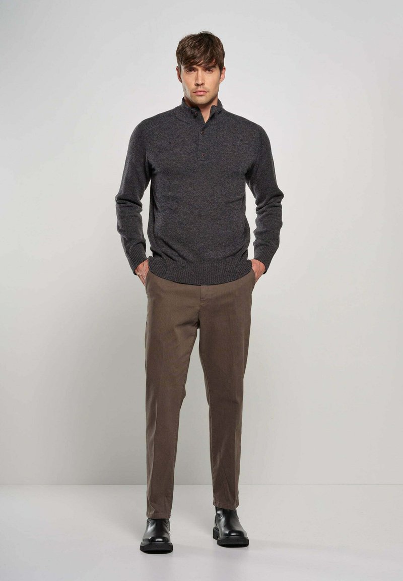 Pullover lavorato a maglia grigio ardesia con colletto alto e un bottone, abbinato a pantaloni chiari a fantasia marrone e stivali neri alla caviglia.