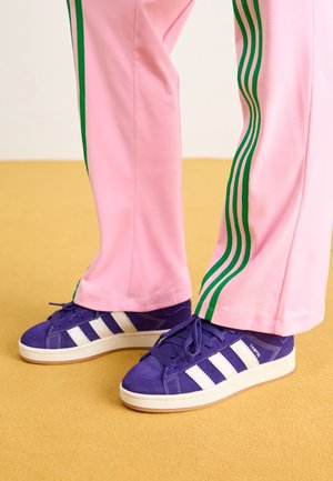Fioletowe trampki Adidas Campus z białymi paskami noszone z różowymi spodniami dresowymi z zielonymi paskami po bokach na żółtej podłodze.