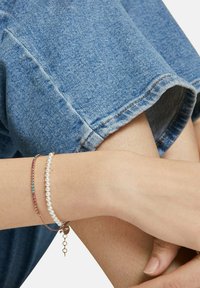 Dünne, geschichtete Armbänder an einem Handgelenk: eines mit kleinen, runden Perlen, ein anderes mit bunten Steinen, versehen mit einem goldenen Verschluss und Kettenakzenten.