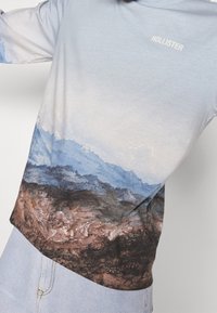 Hellblaues Langarmshirt mit Berglandschaftsprint am Saum, aus weichem Stoff, mit "HOLLISTER" in Weiß bedruckt.