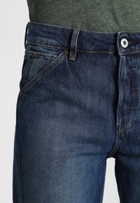Jeans de mezclilla en azul oscuro con una textura suave, que presentan un diseño clásico de cinco bolsillos, costuras en contraste y cierre con botón metálico.