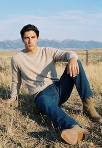 Jeune homme portant un pull beige, un jean bleu et des bottes marron assis sur de l'herbe sèche avec des montagnes en arrière-plan sous un ciel clair.