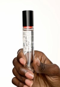 Bouteille de parfum en verre transparent avec un capuchon noir et une accent rouge, ornée de texte imprimé et de motifs, tenue dans une main avec des ongles de teinte claire.