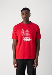 Red cotton t-shirt featuring white graphic print of a sailboat and text. Short sleeves, crew neck, relaxed fit, with smooth texture.