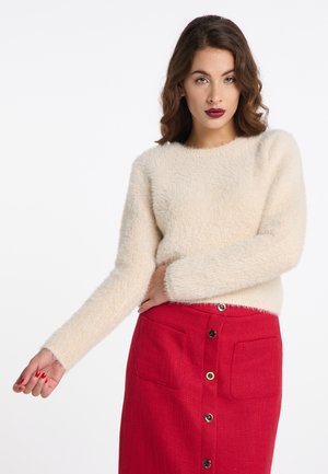 Donna con capelli castani mossi che indossa un maglione crema peloso e una gonna rossa con bottoni, posando su uno sfondo bianco.