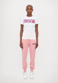 Valge lühikeste varrukatega T-särk, millel on julge roosa "VERSACE JEANS COUTURE" tekst, koos heledate roosade dressipükstega, millel on sobiv logo ning valged spordijalatsid.