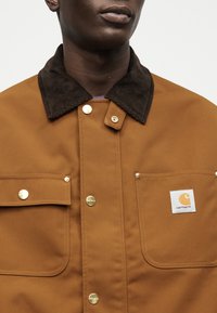 Άτομο με σκουρόχρωμο δέρμα που φοράει καφέ μπουφάν Carhartt με σκούρο γιακά από κορδόνι και χρυσά κουμπιά τύπου σνάπ, εμφανής περιοχή στήθους και λαιμού.