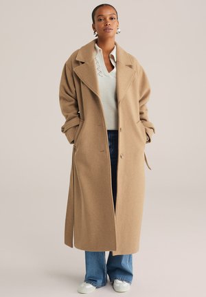 Abrigo largo de lana color camel con solapas amplias, botones de un solo lado y bolsillos profundos, combinado con un suéter ligero y jeans acampanados.