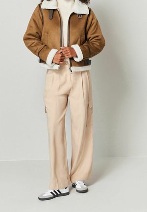 Persona que lleva una chaqueta de piel marrón, pantalones beige de pernera ancha con bolsillos laterales y zapatillas blancas con rayas negras, de pie frente a una pared lisa.