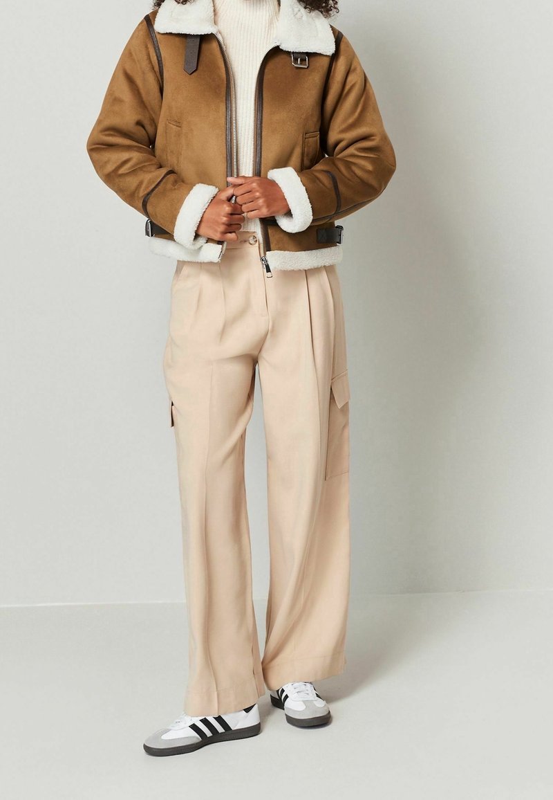 Personne portant une veste en peau retournée marron, un pantalon beige à jambes larges avec des poches latérales et des baskets blanches à rayures noires, debout contre un mur uni.