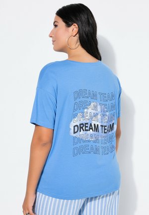 Kobieta z długimi ciemnymi włosami, nosząca duże kolczyki koła, niebieską koszulkę z powtarzającym się napisem "DREAM TEAM" i niebieskie paskowane spodnie, zwrócona bokiem.