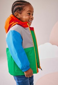 Chaqueta colorida con capucha naranja, cuerpo gris y acentos azules, verdes y rojos; puños elásticos y bolsillos frontales; textura suave.