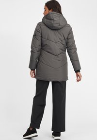 Grå puffjakke med hette, horisontal og diagonal quiltet mønster, lange ermer og elastiske mansjetter, brukt med svarte løse bukser.