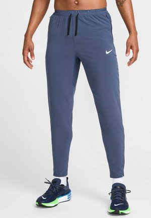 Homme portant un pantalon de course Nike bleu foncé avec taille à cordon et des chaussures de course Nike bleu marine, vertes et blanches, debout, les bras détendus.