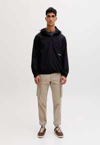 Junger Mann steht, trägt eine schwarze Reißverschlussjacke, beige Cargohosen und braune Turnschuhe vor einem schlichten hellen Hintergrund.