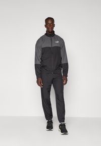 Schwarzes Trainingsanzug mit einem grauen Paneel auf der oberen Hälfte, ausgestattet mit elastischen Bündchen und Bund. Getragen mit schwarzen Laufschuhen und einem dezenten Logo.
