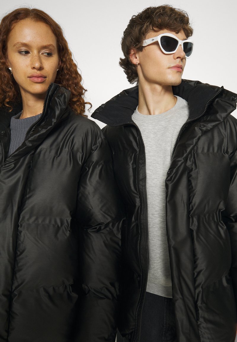 Chaquetas de plumas negras con un acabado brillante, que cuentan con cuellos altos y cierres a presión. El modelo a la izquierda tiene el pelo rizado y rojo; el otro lleva gafas de sol blancas.