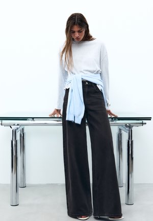 Femme portant un pull gris clair, un jean noir à jambes larges et un pull bleu clair noué autour de la taille, debout près d'une table en verre avec des pieds chromés.