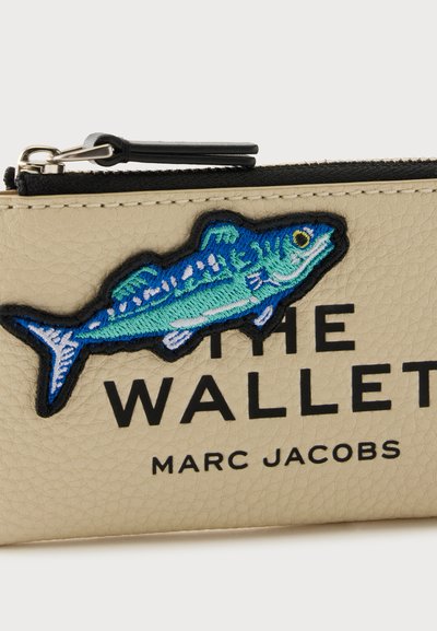 Μπεζ πορτοφόλι Marc Jacobs με μπλε κεντημένο έμβλημα ψαριού, μαύρο φερμουάρ και έντονα μαύρα γράμματα που γράφουν THE WALLET και MARC JACOBS.