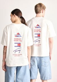 T-shirts brancas de manga curta apresentam um logótipo gráfico nas costas, com elementos de design em vermelho, azul e roxo e texto. Usadas com calções azul claro.