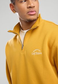 Yellow pullover, quarter-zip design, made of soft fabric. Features white embroidered text "Members Only Golf Riders." Textured finish.