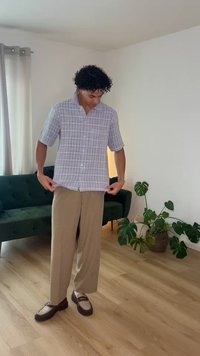 Άνδρας που ρυθμίζει γκρι ανοιχτό καρό πουκάμισο, φοράει μπεζ παντελόνι και μοκασίνια, στέκεται σε ξύλινο πάτωμα κοντά σε πράσινο καναπέ και γλαστράκι.