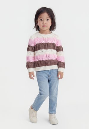 Malé dítě stojící s rukama u těla, oblečené v pruhovaném růžovém, hnědém a bílém pleteném svetru, modrých džínách a bílých teniskách.