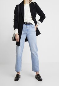 Czarny długi płaszcz, kremowy sweter z dzianiny, jasnoniebieskie dżinsy o prostym kroju, czarne mokasyny ze złotym okuciem i czarny pasek.