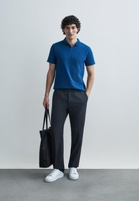 Zils polo krekls ar apkakli, kopā ar tumši pelēkām elastīga vidukļa biksēm. Modelis tur melnu lielveikala somu, valkājot baltas sporta kurpes.