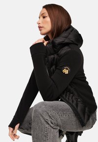 Zwarte puffer vest met een capuchon, getailleerd ontwerp en gouden logo accent. Draagt over een zwart langemouwen top, gecombineerd met grijze textuurpants.