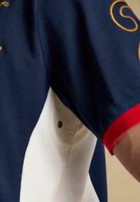 Manche de polo bleu marine et blanc avec bordure rouge, motif brodé jaune près de l'épaule, et un oeillet de ventilation métallique dans le tissu blanc.