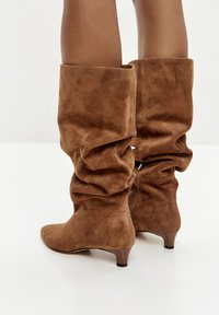 Bruine nubuck kniehoge laarzen met een slonzig ontwerp, spitse neus en een lage hak. Zichtbare stiksels en een ritsdetail aan de zijkant.