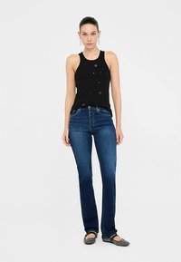 Must tanki tipus lillekaunistustega, kombineeritud tumeliste siniste sirgete pükstega. Modell kannab musti lamejalanõusid, seisab ühevärvilise tausta ees.