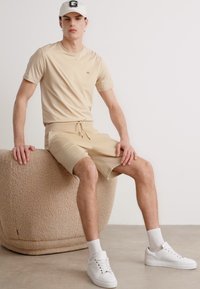 Beige pamut póló és hozzá illő, zsinórral ellátott sort, fehér sportcipővel párosítva. Egyén egy textúrált bézs széken ül.