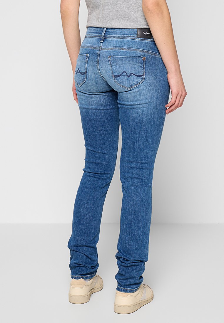 Person in engen, blauen Jeans mit bestickten Gesäßtaschen und beigen Sneakers, vor einem schlichten weißen Hintergrund stehend.