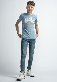 Camiseta de algodón azul claro con gráfico de motocicleta y texto naranja, combinada con jeans de lavado medio y zapatillas blancas.