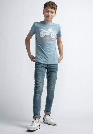 Lyseblå bomulds T-shirt med motorcykelgrafik og orange tekst, parret med medium-vasket denimjeans og hvide sneakers.